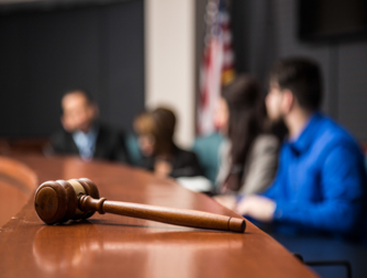A gavel laying on a brown table in a court room with people sitting around the table.