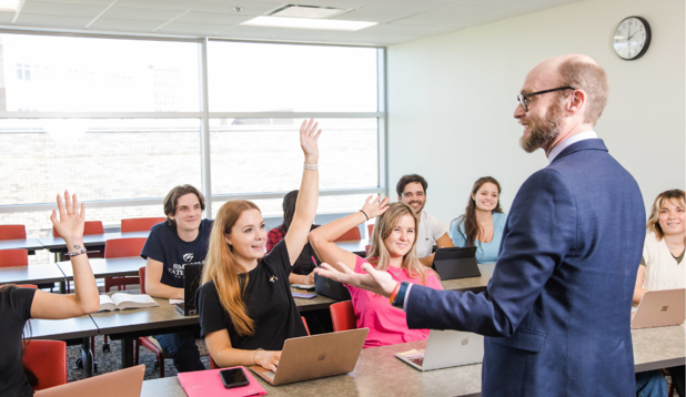 Professor teaching in a classroom to students and the students are raising their hands to ask questions
