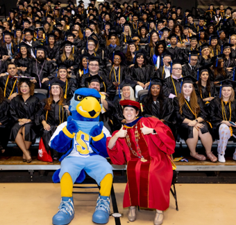 Rally Raider and Dr. Lorenz sitting in the auditorium in front graduation students, showing a thumbs up symbol to the camera