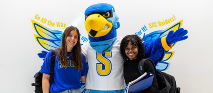 Rally Raider standing with two students with a wing design in the background.