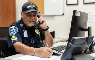 A safety officer sitting at an office desk in front of a computer, talking on the phone, writing notes on a piece of paper.