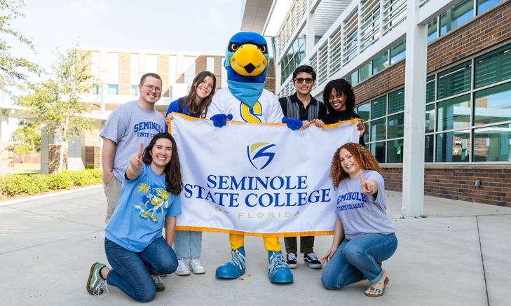Rally the Raider and six SSC employees posing with a Seminole State College of Florida banner outside at the Lee Campus at Oviedo.